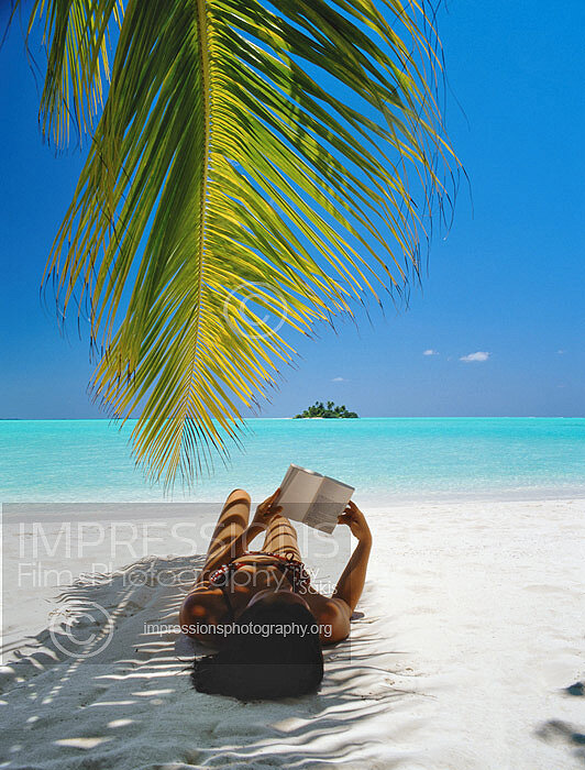 Maldives, woman lying under shade of palm tree on beach, reading