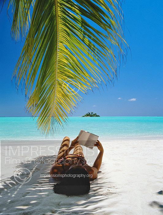 Maldives, woman lying under shade of palm tree on beach, reading book